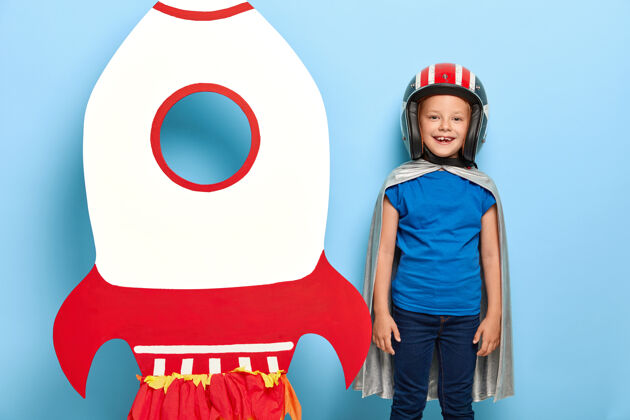 探索快乐的小孩戴着头盔和灰色斗篷 站在纸飞船火箭旁 想在太空中飞行服装头盔服装