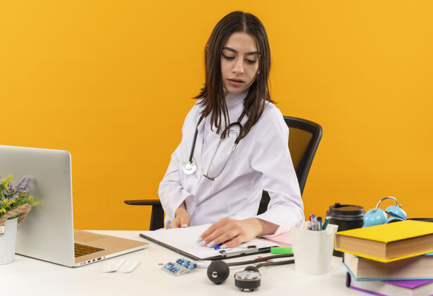 商务忙碌的年轻女医生 穿着白大褂 手持听诊器 坐在桌旁 手提电脑和文件放在橙色的墙上医疗保健坐着女人
