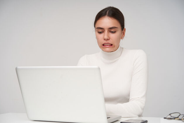 头发困惑的年轻漂亮的黑发女性坐在白色墙壁上的桌子旁 穿着白色针织圆领衫 拿着现代化的笔记本电脑 一边看着屏幕一边做鬼脸现代工作编织