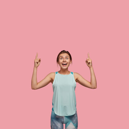 时尚积极的欧洲女人 表情愉快 食指朝上 穿着休闲背心和紧身裤 模特们站在粉红色的墙上广告概念看天花板大笑广告休闲