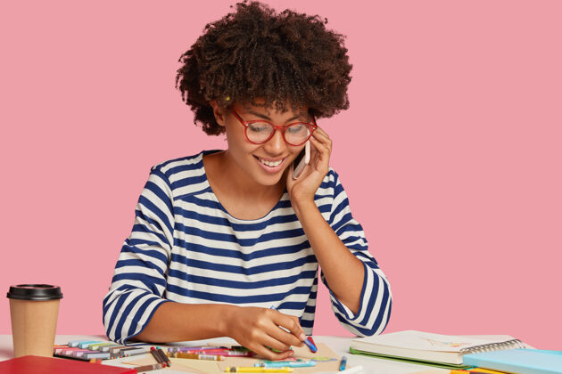 日记快乐的黑人女士喜欢画画 在白纸上作画 戴眼镜 打电话 微笑着谈论一些愉快的事情 孤立在粉红色的墙上饮料女商人工作