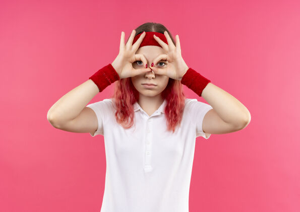 运动员戴着头巾的年轻运动女性用望远镜握着手指 手指穿过手指站在粉红色的墙上头带人市民