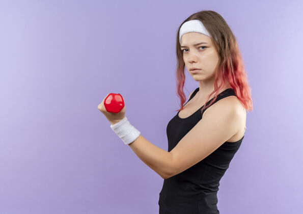 女人身穿运动服的年轻健身女士正脸站在紫色的墙上用哑铃做练习运动站脸