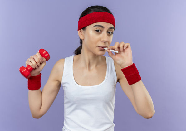 人身穿运动服的年轻健身女士举着哑铃 手里拿着香烟 站在紫色的墙上抽烟吸烟站健身