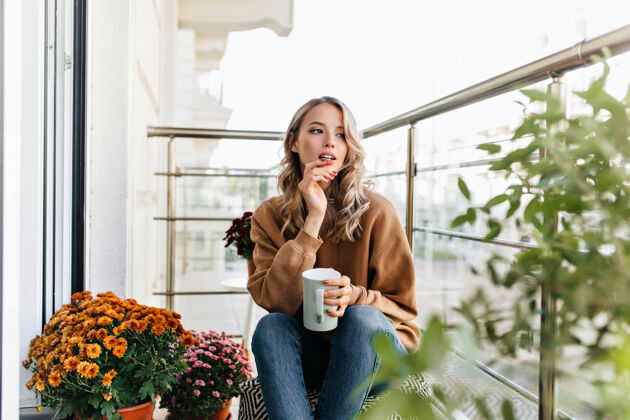 模特浪漫的女孩坐在鲜花旁 端着一杯咖啡无忧无虑的年轻女子在想什么欢笑女性寒冷