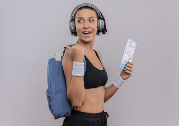 拿着穿着运动服的年轻健身女士头戴耳机背着背包拿着机票看着一边用拇指指着后面站在白色的墙上票女运动装