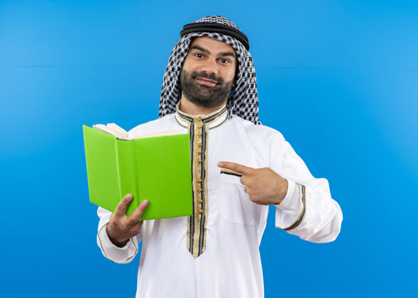 商务人士身着传统服饰的阿拉伯商人手拿一本打开的书 用手指着书 脸上带着微笑站在蓝色的墙上握指穿
