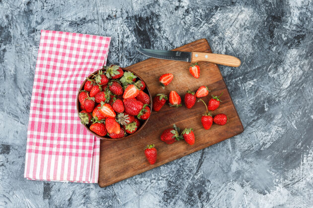 食物把一碗草莓和一把刀平放在木砧板上 用红格子桌布铺在深蓝色大理石表面水平平板切片果酱