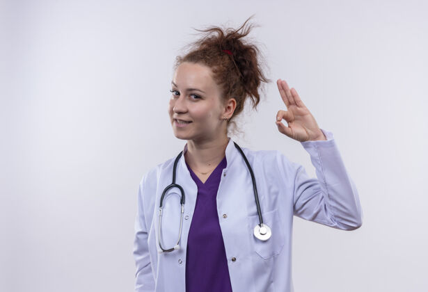 穿上年轻的女医生穿着白大褂 手持听诊器 微笑着 自信地站在白墙上做着“没事”的手势听诊器自信女人