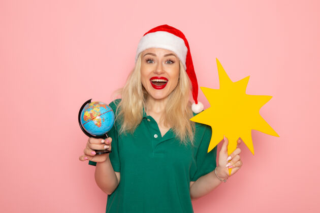美丽正面图年轻女性手持地球仪和粉色墙上的黄色人物模型圣诞节假期照片新年色彩身材圣诞节风景