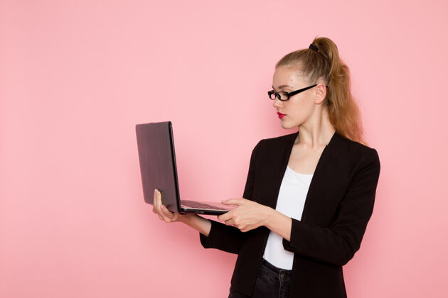 笔记本电脑穿着黑色夹克的女上班族在粉色墙上使用笔记本电脑的前视图女性严格工作