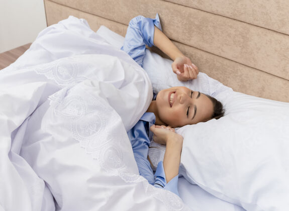 醒来穿着蓝色睡衣的年轻漂亮女人躺在床上 靠着柔软的枕头 醒来时在卧室的室内灯光背景下伸展双手睡衣柔软床