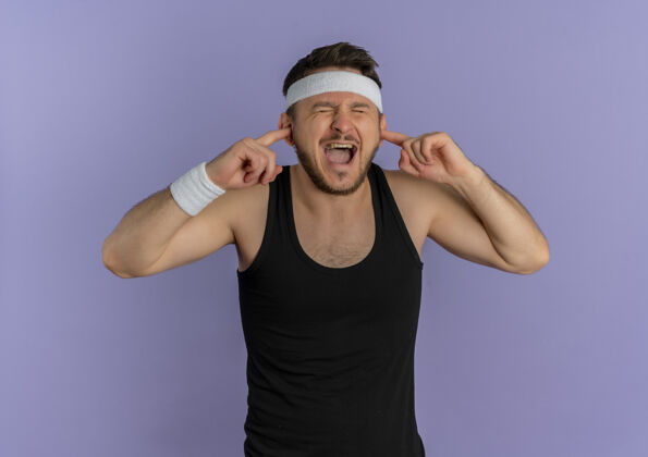头带戴着头巾的年轻健身男子站在紫色的墙上用手指捂住耳朵 脸上露出恼怒的表情男人运动员健康