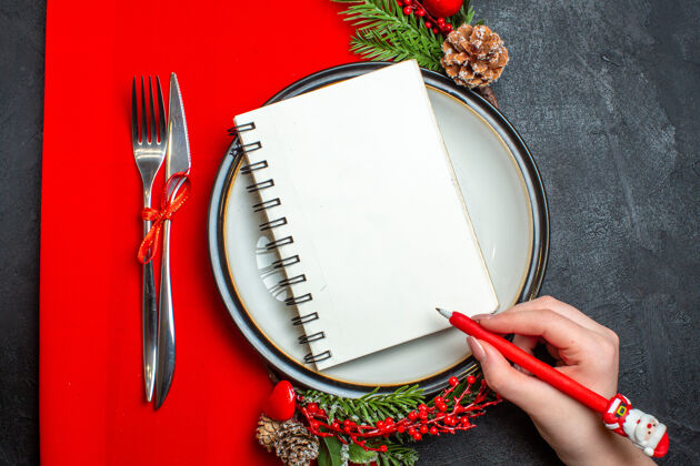 钢笔手拿笔放在盘子上的螺旋形笔记本上的俯视图 装饰配件杉木树枝和餐具放在红色餐巾上盘子螺旋餐具