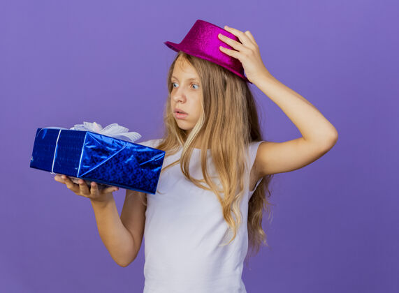 盒子漂亮的小女孩戴着节日礼帽拿着礼盒看着它兴致勃勃 生日派对的概念站在紫色的背景上紫色站小