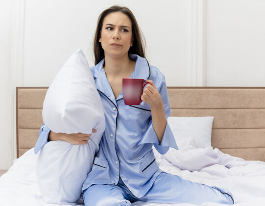 女人穿着蓝色睡衣的年轻美女坐在床上 带着枕头和一杯咖啡 在浅色背景下看着卧室里不开心的脸室内床年轻