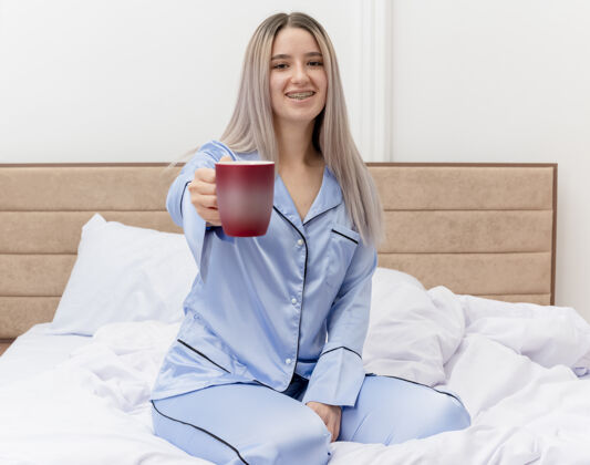 杯子穿着蓝色睡衣的年轻美女坐在床上 端着一杯咖啡 在卧室的室内灯光背景下欢快地微笑着睡衣女士提供
