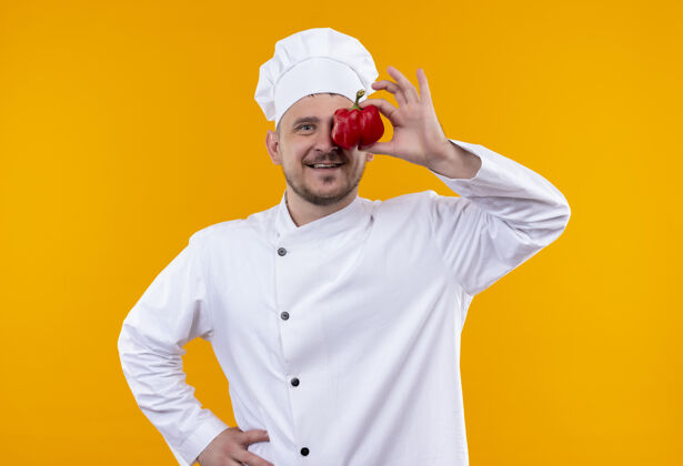 胡椒面带微笑的年轻帅哥厨师身着厨师制服把辣椒放在眼睛上孤立的橙色空间厨师眼睛放