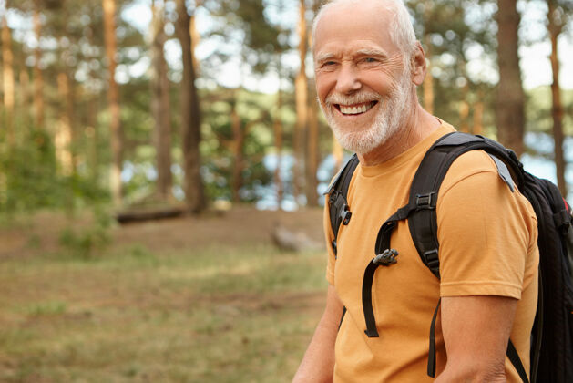 背包快乐精力充沛的退休老人背着黑色的背包 笑容满面 在阳光明媚的秋日里享受着在树林里徒步旅行的乐趣野外拍摄的留着胡子的老人在森林里散步老人男人乡村