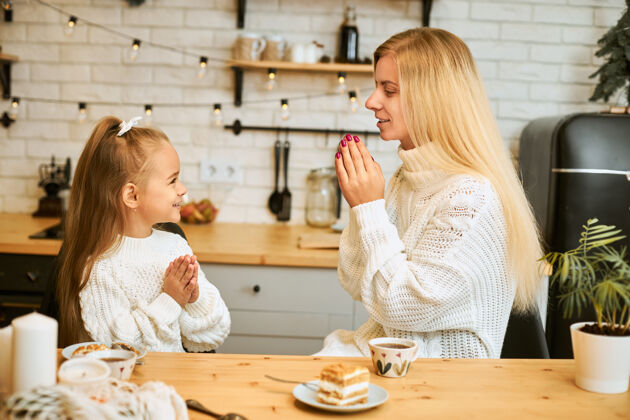 享受侧视图：穿着白毛衣的年轻白种人美女在饭前说优雅的话 她和她的小女儿坐在厨房的桌子旁 双手紧握在一起 去吃蛋糕和喝茶家庭女儿孩子