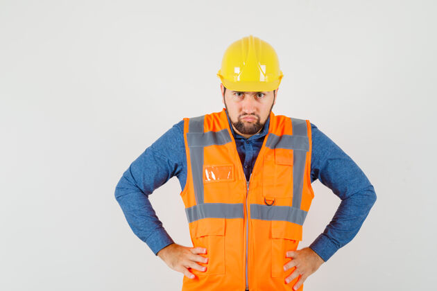 工人年轻的建设者站着 手放在腰上 穿着衬衫 背心 戴着头盔 看上去很紧张 正对着前方安全帽工具工匠
