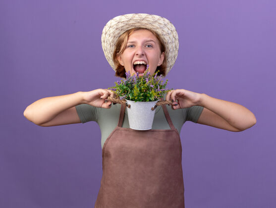 园丁愤怒的年轻斯拉夫女园丁戴着园艺帽 手里拿着花盆里的花 隔离在紫色的墙上 留着复制空间花帽子年轻