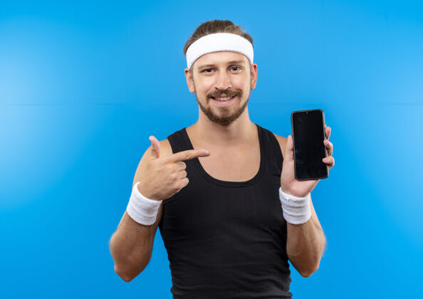 微笑面带微笑的年轻帅气的运动型男人戴着头带和腕带拿着手机指着蓝色的空间手持年轻帅气