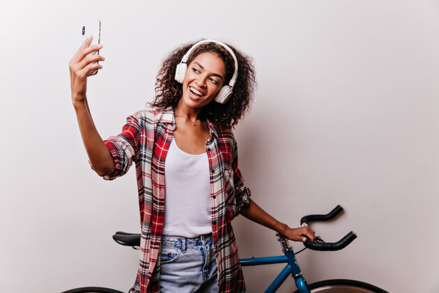 自行车喜欢卷发的女孩骑自行车自拍时尚的非洲女人在白色地板上欣赏音乐的室内镜头积极黑色休闲
