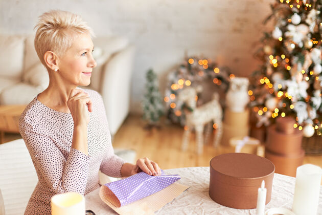 幸福欢快成熟的欧洲短发女性准备新年或圣诞节庆典的室内镜头 餐桌上摆着礼品纸坐在客厅里 有着沉思沉思的神情 微笑着剪刀包装包装