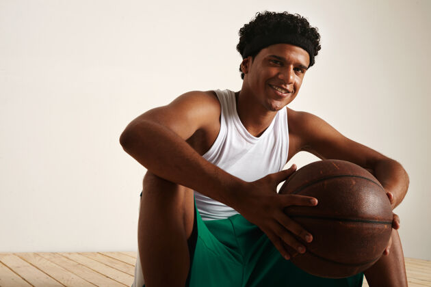 Positive坐着微笑友好的非洲裔美国篮球运动员与非洲穿着白色和绿色制服举行一个棕色皮球LeatherGameOutdoor