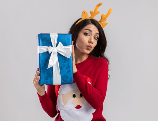 圣诞老人自信的年轻漂亮女孩戴着驯鹿鹿角头带和圣诞老人毛衣拿着圣诞礼物包撅着嘴唇看着年轻鹿角包装