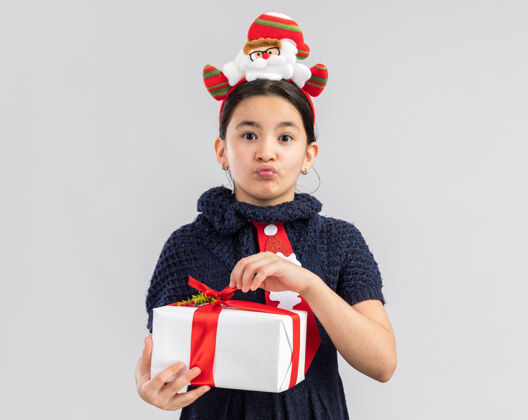 新的穿着针织连衣裙的小女孩头上系着一条红色领带 头上戴着一个滑稽的圣诞边 手里拿着圣诞礼物 看上去很困惑小红色困惑