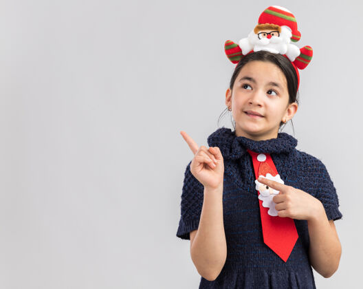 快乐快乐的小女孩穿着针织连衣裙 头上系着一条红色领带 戴着有趣的圣诞戒指 看着一边 用食指指着复印区空间女孩新