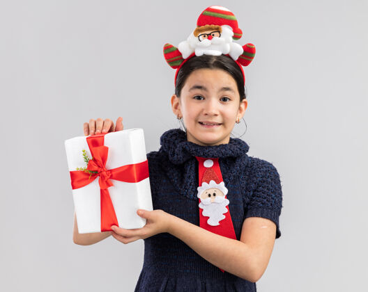 编织快乐快乐的小女孩 穿着针织连衣裙 头上系着一条红色领带 头上戴着一个有趣的圣诞圈 手里拿着圣诞礼物 脸上挂着微笑抱着领带搞笑