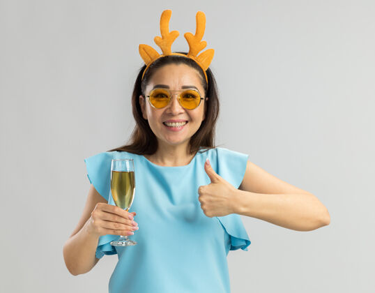 新年身穿蓝色上衣的年轻女子 戴着滑稽的鹿角边 戴着黄色的玻璃杯 手里拿着一杯香槟 脸上露出愉快的笑容 竖起大拇指角快乐看着