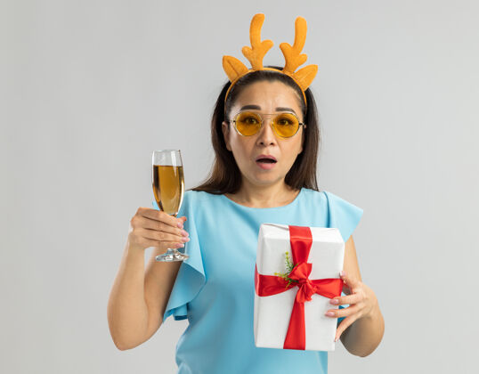 圣诞身穿蓝色上衣的年轻女子戴着滑稽的鹿角边 戴着黄色的眼镜 手里拿着一杯香槟和圣诞礼物 看上去既焦虑又困惑顶快乐黄色