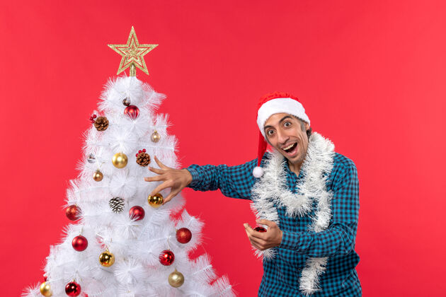 圣诞一个戴着圣诞老人帽子的年轻人 穿着一件蓝色的衬衫 把圣诞树装饰成令人惊讶的红色衬衫圣诞老人装饰