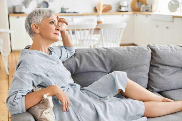沙发迷人时尚的欧洲女性退休者的侧视图 穿着优雅的礼服 光着脚坐在舒适的沙发上 有着深思熟虑的神情 白日梦人 退休 成熟的年龄和生活方式平静沙发女人