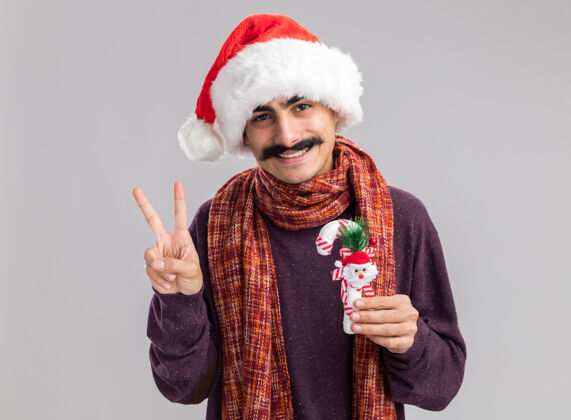 新快乐和积极的年轻胡子男子戴着圣诞老人帽子和温暖的围巾在他的脖子上举行圣诞糖果手杖微笑显示v-标志围巾圣诞快乐脖子