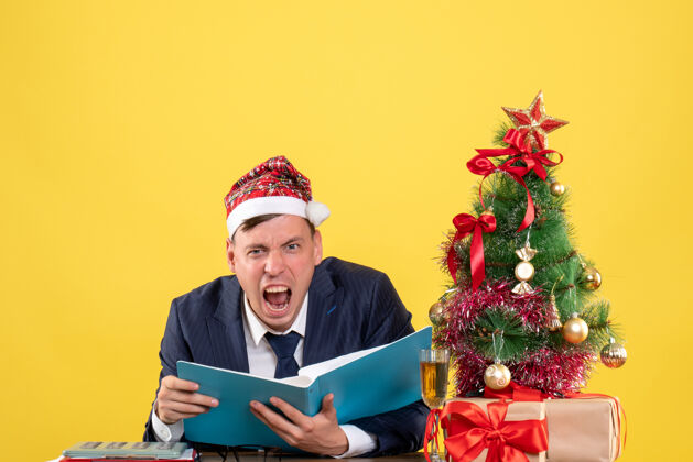 电脑前视图：一个喊着的商人坐在圣诞树旁的桌子旁 黄色的礼物人商人树