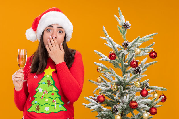 新带着圣诞帽的焦虑的白人女孩把手放在嘴上 拿着一杯香槟 站在橙色背景下的圣诞树旁香槟杯子圣诞老人