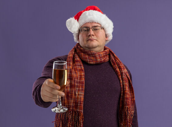 自信自信的成年男子戴着眼镜 戴着圣诞帽 脖子上围着围巾 向镜头伸出香槟酒杯 看着隔离在紫色背景上的镜头香槟圣诞老人眼镜