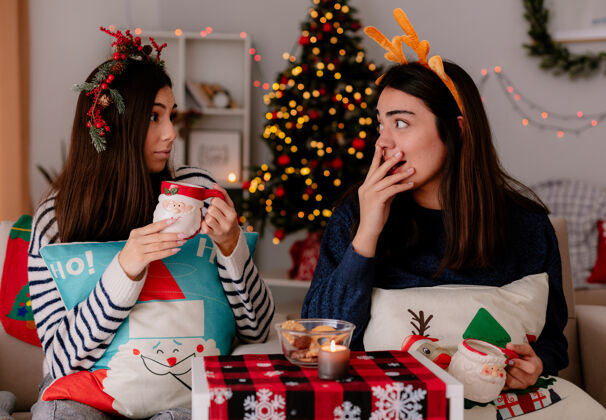 扶手椅带着冬青花环和驯鹿头带的漂亮女孩们拿着杯子 坐在扶手椅上互相看着对方 在家里享受圣诞节时光举行圣诞快乐每个