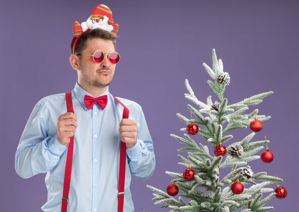 站立严肃的年轻人穿着吊带领结 戴着圣诞老人和红眼镜 站在圣诞树旁 在紫色的墙上自信地看着男人圣诞老人蝴蝶结