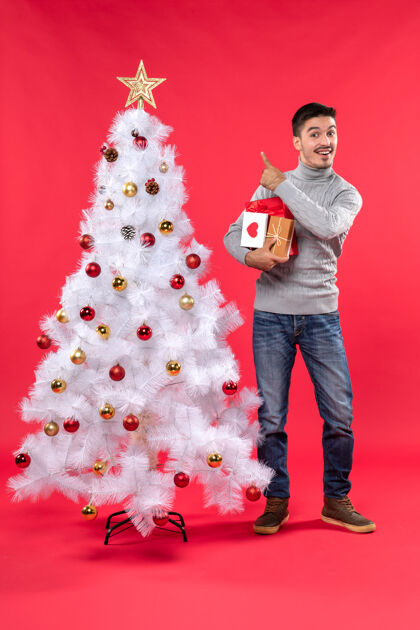 圣诞树英俊的年轻人站在装饰过的白色新年树旁 手里拿着礼物 指着红色的东西年轻人帅哥礼物