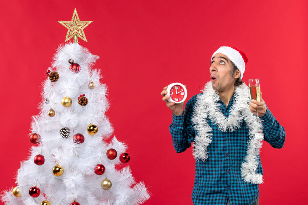 圣诞一个戴着圣诞老人帽子 拿着一杯葡萄酒和一个挂钟 站在红色圣诞树旁的年轻人吓了一跳杯子站着圣诞老人