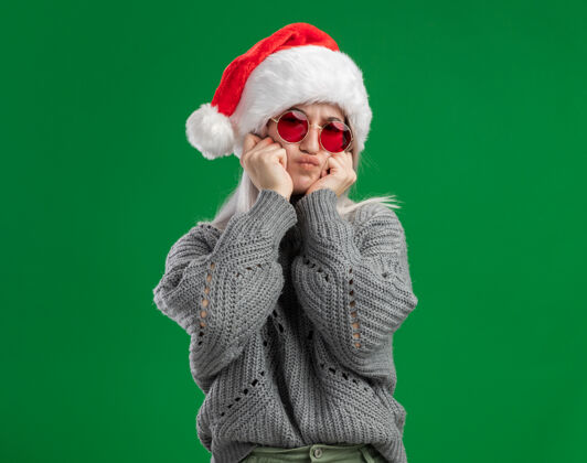 圣诞老人年轻的金发女郎穿着冬衣 戴着圣诞帽 戴着红眼镜 看着摄像机做鬼脸 站在绿色的背景下玩得很开心圣诞眼镜有