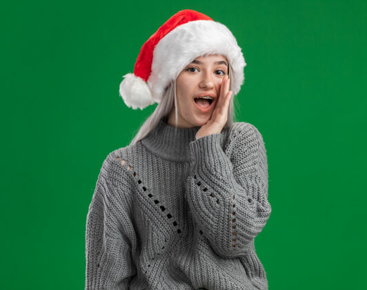 近年轻的金发女郎穿着冬季毛衣 戴着圣诞帽 站在绿色的背景下 用手在嘴边低语着秘密金发帽子毛衣
