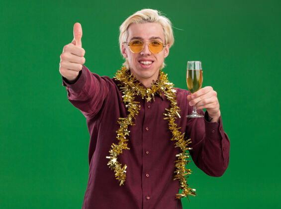 玻璃杯微笑着的年轻金发男子戴着眼镜 脖子上戴着金箔花环 手里拿着一杯香槟 在绿色的墙上孤立地竖起大拇指年轻人周围香槟