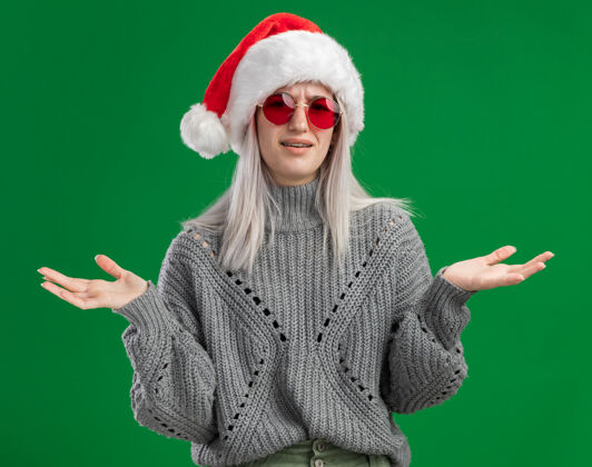 站着年轻的金发女郎穿着冬衣 戴着圣诞帽 戴着红眼镜 困惑地看着镜头 双臂伸向两侧 站在绿色的背景上手臂快乐圣诞老人